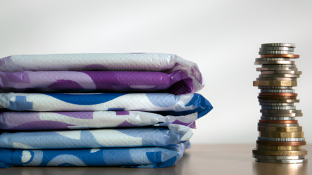Une pile de protections hygiéniques jetables emballées à côté d'une colonne de pièces de monnaie, illustrant le coût des protections hygiéniques et le coût important de ces protections menstruelles.