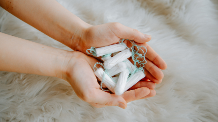 Gros plan de deux mains jointes tenant plusieurs tampons blancs sur un fond de fourrure synthétique blanche, illustrant la distribution de protections menstruelles et l'accès à une solution hygiénique dans le cadre de la lutte contre la précarité menstruelle.