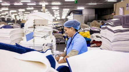 Une couturière professionnelle en plein travail de confection textile dans un atelier, entourée de piles de tissus blancs, illustrant la fabrication de qualité de nos solutions contre la précarité menstruelle et de nos protections hygiéniques durables.